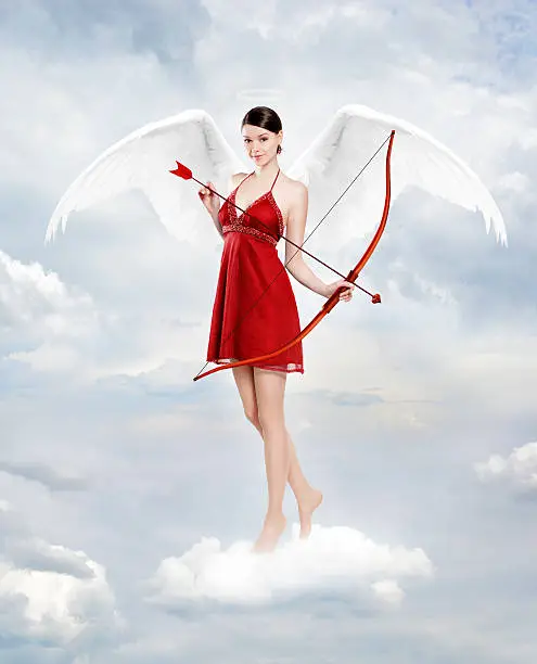 Gorgeous young brunette woman as cupid angel in clouds with white wings, bow and arow