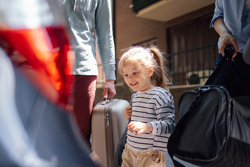 Close up shot of a cute little girl walking with her parents to the car. They are carrying bags while she is going in front of them excited to go on the vacation with them.