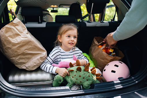 Portrait of a cute playful girl sitting in the boot of the car smiling and looking away. An unrecognizable man is packing bags in the car. They are getting ready to go on a trip or a day in nature or picnic.