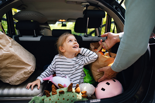 Portrait of a cute playful girl sitting in the boot of the car smiling and looking up. An unrecognizable man is packing bags in the car. They are getting ready to go on a trip or a day in nature or picnic.