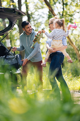 Happy couple with their daughter standing in the forest next to their car. The father is taking a bag out of the boot while the mother is holding their child. They are looking at each other and smiling.