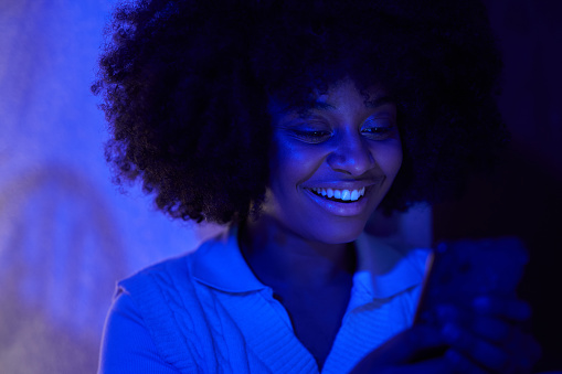 Front view of a young African American woman smiling while using her cell phone illuminated with a blue light.