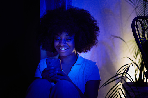 Portrait of a young African American woman using her cell phone sitting on the floor and leaning against the wall illuminated with a blue light,