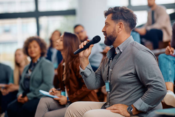 istock Business seminar attendee talking on microphone while sitting in the audience at convention center. 1604032413