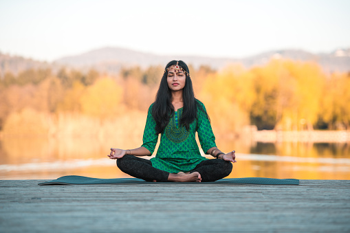 Full length image of an  attractive young Indian female, her long hair falling on her shoulders, sitting on a yoga mat and meditating in lotus position. Eyes closed, wearing a traditional Indian hair  decoration and comfortable clothes. Dawn breaking on the blurred trees and water in the background.