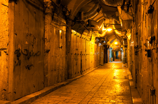 Shuttered shops at night on Beit HaBad Street in the Muslim Quarter of Jerusalem's Old City. The street is a major commercial street during the day. In the distance are people (blurred motion).