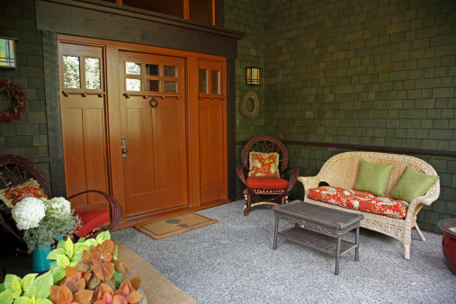  Outdoor porch of Shingle Style Craftsman Bungalow.  Bent willow chairs and rattan sofa and table.  Flowers and plants in pots with floral fabrics on furniture.  Custom craftsman Mission style lights with wooden door.