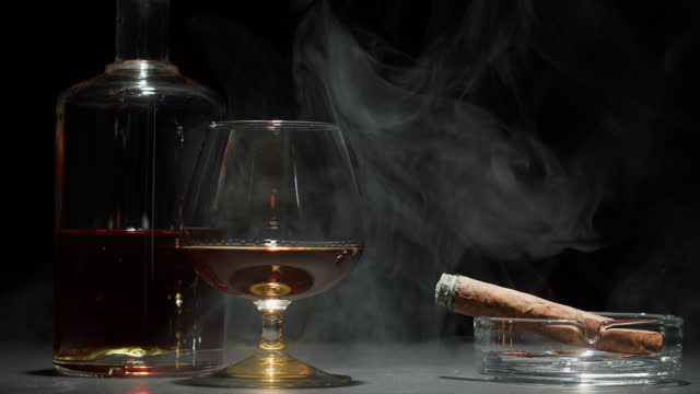 Brandy or whiskey and cigar close-up. Smoking cigarette and drinking luxury cognac on black background. Alcohol amber drink, rum, liqour beverage in glass.