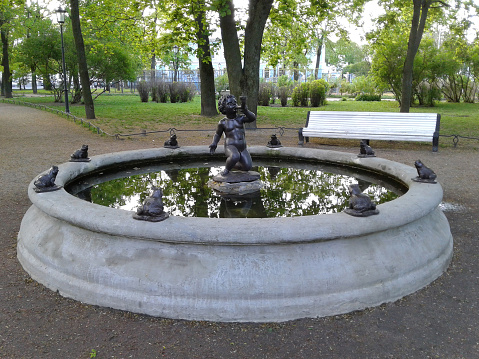 Fountain in Nikolsky square. Sculpture of a boy with frogs. The boy hums into the horn, around him there are 8 frogs. Reflection of trees in the water. Summer day morning. The fountain is off. Silence, empty bench. Places to stay in the city center.