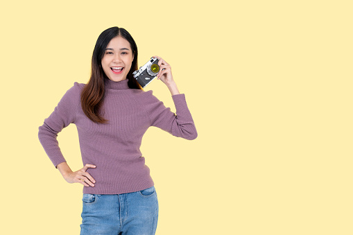 A happy and beautiful Asian woman is holding her vintage film camera, standing against an isolated yellow background. traveler, tourist, hobby and lifestyle