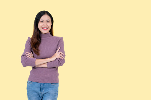 A portrait of an attractive and smiling Asian woman in casual clothes stands with her arms crossed against an isolated yellow background.