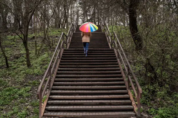 Photo of young woman in a beige coat and rain boots stands in forest or park and holds colorful bright rainbow umbrella. Autumn weather in city, girl is walking on a wooden stairs in the park. High stairway