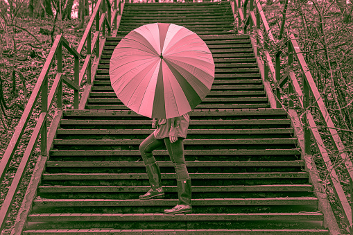Photo in pink tones. Young woman in a coat and boots stands on stairs and holds bright umbrella. Total pink