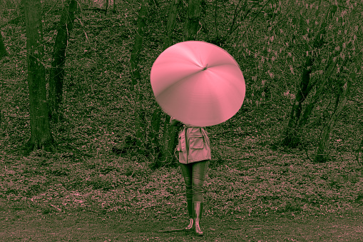 Photo in pink tones. Young woman in a coat and boots stands in forest or park and holds bright umbrella. Girl spins umbrella for blur effect. Slow shutter speed. Autumn weather in city. Total pink