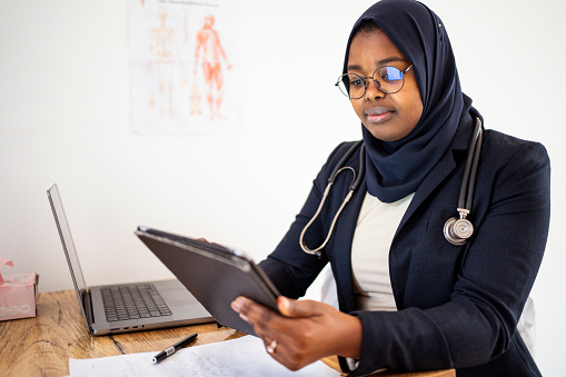 Young muslim female doctor wearing a hijab using a digital tablet at a desk in the office of her medical practice