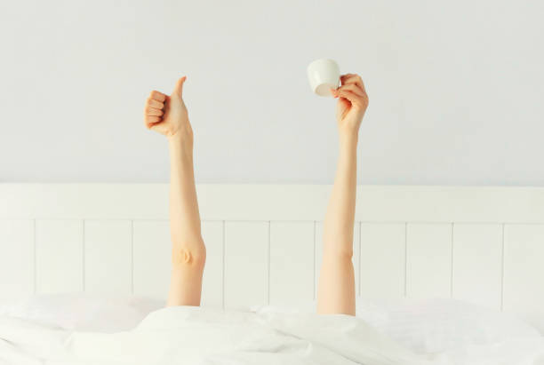 Cheerful lazy woman waking up after sleeping lying in soft comfortable bed showing empty cup coffee stretching her hands up from under the blanket in white bedroom stock photo