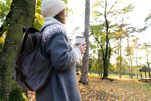Woman traveler with backpack is holding a cup of hot drink in autumn park.