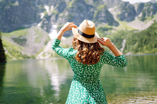 Young tourist woman in bright green dress and hat on picturesque background of mountains and turquoise mountain lake. Happy woman enjoys mountain views. Concept of tourism, vacation.