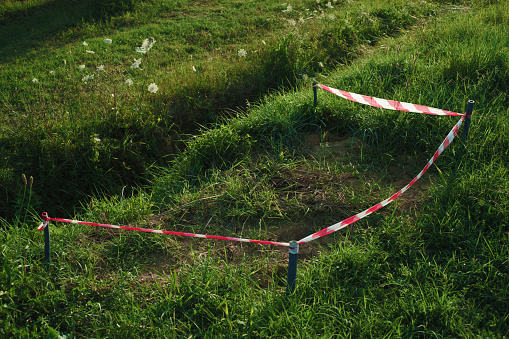 grassy field with a red and white striped tape cordoning off a small area. The tape is attached to two blue poles on either side. The area cordoned off appears to be a small pit or depression in the ground