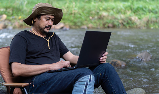 Mature worker Asian man sit down on camping chair and work on the laptop near the stream in the middle of park outdoors forest woods alone.