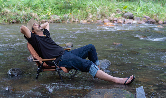 A happy man with hiking hat resting on a camping chair in the stream in the forest and enjoys the surrounding nature on a sunny day. Outdoors llifestyle concept.