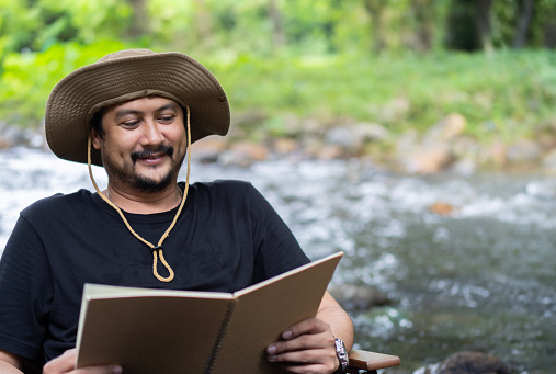 Portrait of happy alone Asian man sitting in chair near the stream on country and enjoying forest view while reading a book. Reading or education concept image