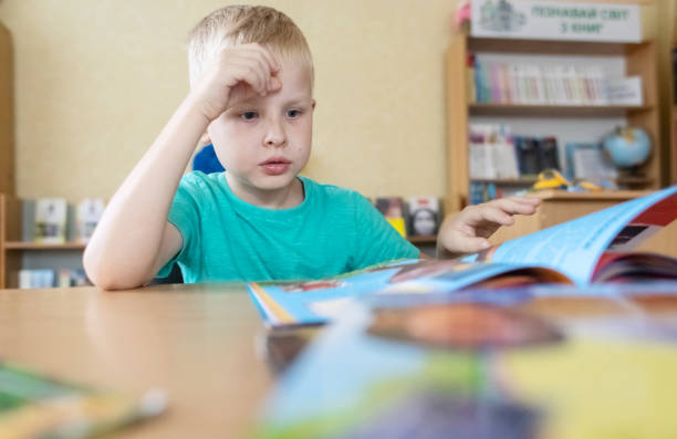 istock Boy in library. Seven year Ukrainian guy is sitting at a table in library and reading book. Concept of normal learning, thirst for knowledge, desire to learn new things. Blurred background and letters 1605017245