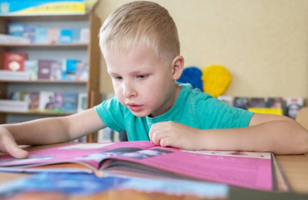 istock Boy in library. Seven year Ukrainian guy is sitting at a table in library and reading book. Concept of normal learning, thirst for knowledge, desire to learn new things. Blurred background and letters 1605017793