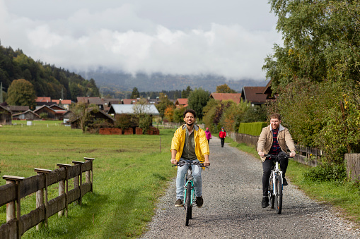 A same sex couple riding their mountain bikes on a gravel path surrounded by wooden huts and greenery while on holiday in Garmisch-Partenkirchen, Germany. They are riding alongside each other while they bond and smile together.
