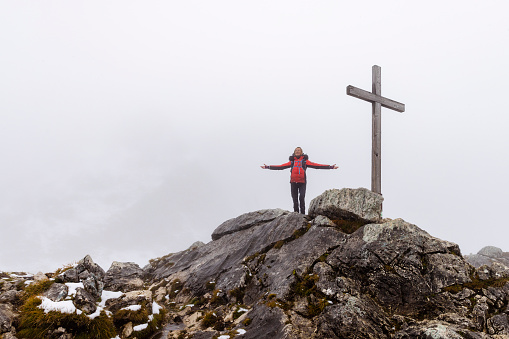 A mature woman wearing a winter coat, standing on a rocky mountain while in Garmisch-Partenkirchen, Germany. She has her arms outstretched and is enjoying the fresh around her while standing next to a religious cross landmark.