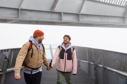 A same sex couple walking on a mountain viewpoint after getting a cable car up to it while on holiday in Garmisch-Partenkirchen, Germany. They are holding hands and walking on a metal walkway after walking to the end of it to look at the view which is not visible due to the fog. One man is looking over his shoulder at his partner and smiling.