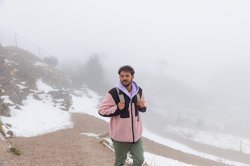 A mid adult man wearing a coat, walking on a footpath on a snowy mountain while on holiday in Garmisch-Partenkirchen, Germany. He is heading to the mountain peak and the view is low visibility due to the fog and snow around him.