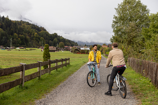A same sex couple taking a break from riding their mountain bikes while on holiday in Garmisch-Partenkirchen, Germany. They are sitting on their bikes on a gravel path surrounded by greenery, mountains and a village while they talk and bond with each other.
