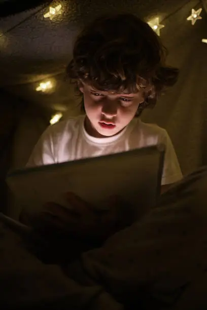 Concentrated talented boy drawing in sketchbook while sitting under blanket construction with glowing garland in dark room at night time