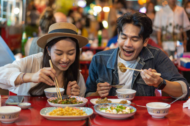 Young Asian couple traveler tourists eating Thai street food together in China town night market in Bangkok in Thailand - people traveling enjoying food culture concept stock photo
