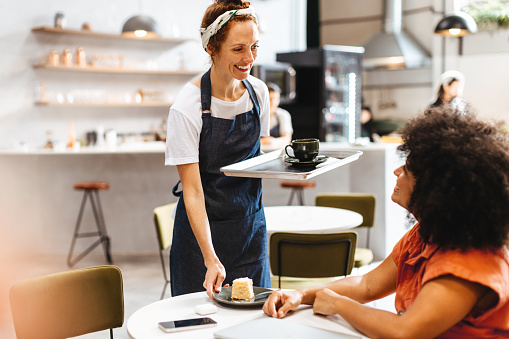 Female server at a restaurant greets her customer with a smile and brings the food on a tray. Friendly female barista serves a customer a cup of coffee along with a slice of cake.