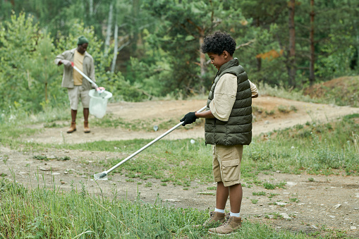 Little boy cleaning the ground with stick together with his dad in background outdoors