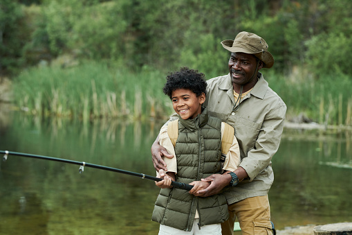 Loving father teaching his smiling young son how to fish with a rod during a vacation together at a scenic lake on a summer afternoon