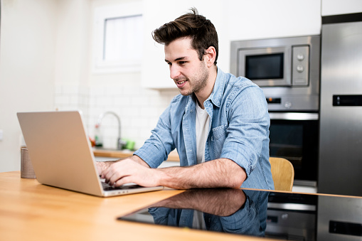 Handsome young man working with a laptop and smiling at the kitchen with a cup of tea - Cheerful, success and business concept