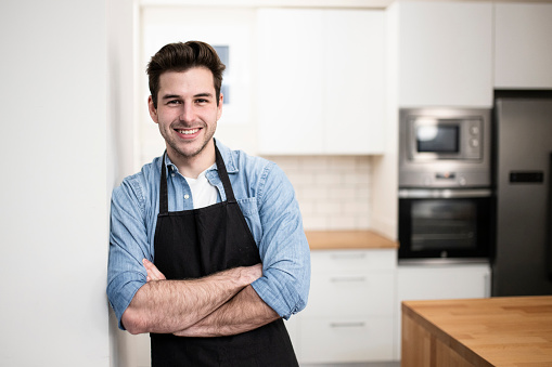 handsome young man smiling in the kitchen wearing an apron - Portrait of a cheerful confident caucasian young man looking at camera with arms crossed . High quality photo