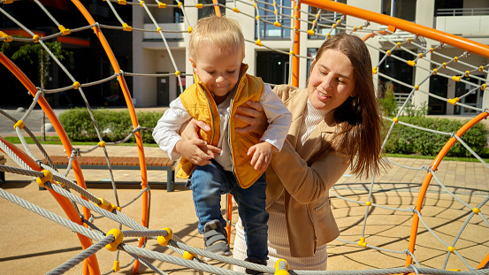 Young mother supporting her baby son balancing on the playground. Children playing outdoor, kids outside, summer holiday and vacation