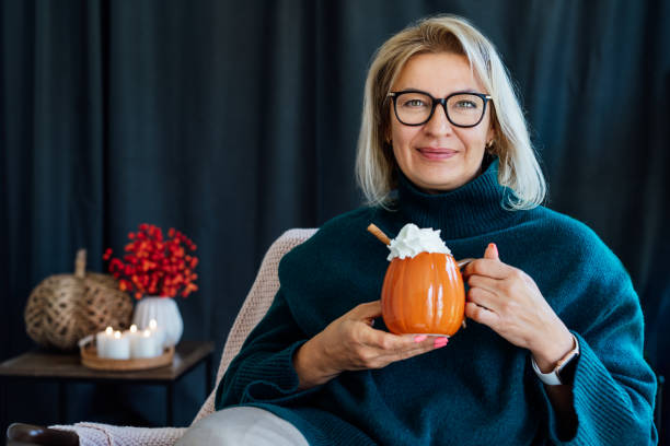 Smiling middle aged woman relaxing with pumpkin shaped cup of hot drink with whipped cream and cinnamon stick with fall mood composition for hygge home decor on background. Cozy autumn holidays mujer sonriente de mediana edad relajándose con una taza de bebida caliente en forma de calabaza con crema batida y rama de canela con composición de humor de otoño para la decoración del hogar hygge en el fondo. acogedoras vacaciones de otoño.