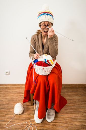 Funny woman with horn rimmed glasses is sitting on a chair and knitting winter caps.