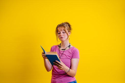 Portrait of a beautiful young woman reading a book while standing in front of a bright yellow background.
