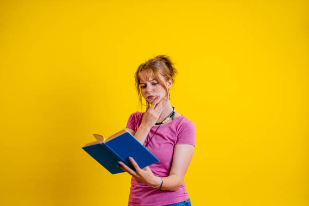 Portrait of a beautiful young woman reading a book while standing in front of a bright yellow background.