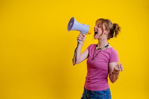 Portrait of a beautiful young woman standing in front of a bright yellow background and talking into the megaphone.