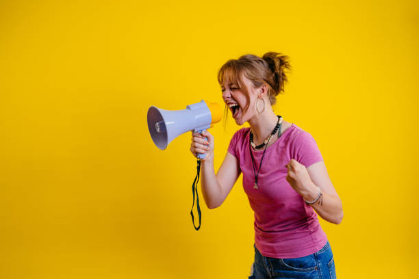 istock Young Woman Talking Into The Megaphone Against A Yellow Background 1605273341