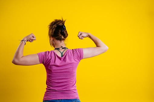 Portrait of a beautiful young woman flexing muscles while standing in front of a bright yellow background.