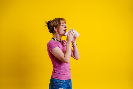 Portrait of a beautiful young woman sneezing into a tissue while standing in front of a bright yellow background.