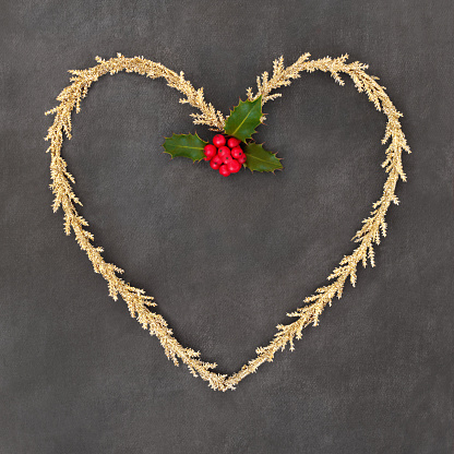 Christmas gold tinsel heart shape wreath with winter holly. Traditional romantic symbol for the festive holiday season on grunge gray background.